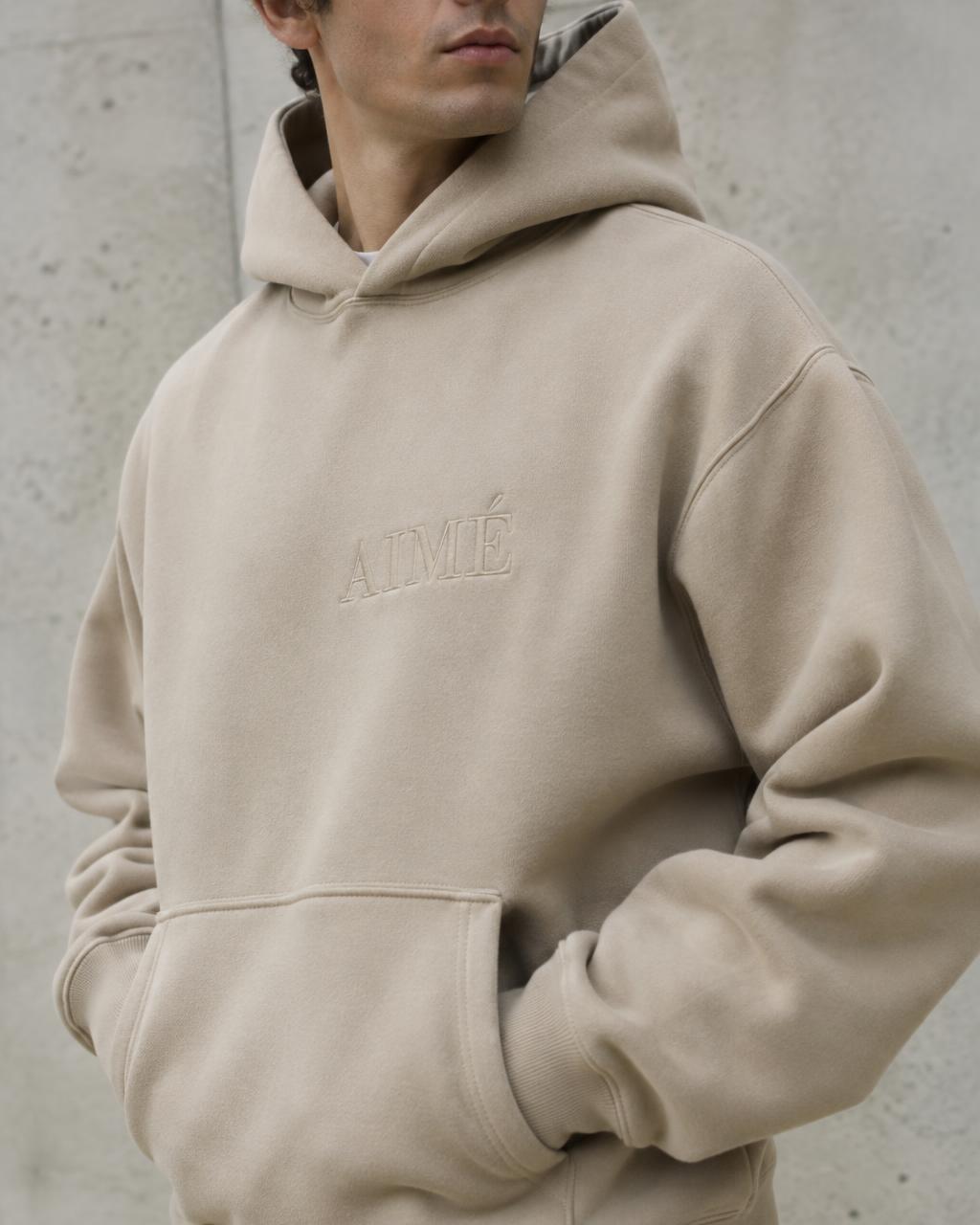 Personne portant un hoodie épais couleur sable avec une broderie ton sur ton sur la poitrine, devant un mur en béton clair